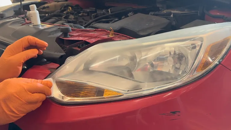 An orange-gloved hand wipes a small white applicator across the lower corner of the headlight lens.