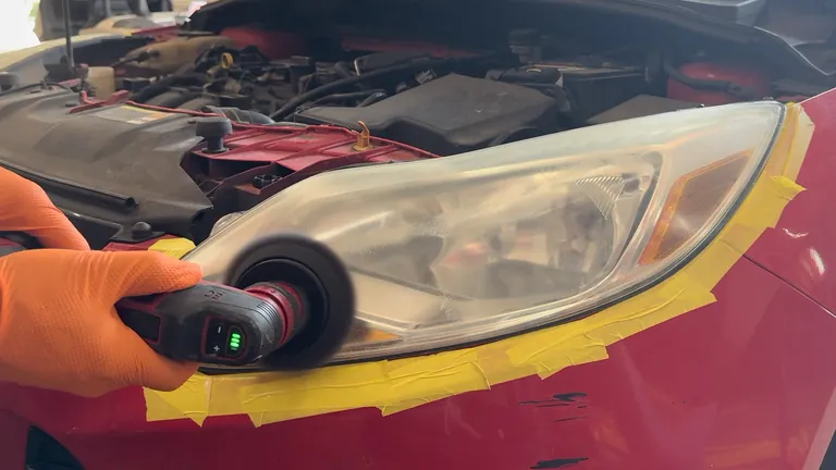 Orange-gloved hands guide a compact power polisher along the lower part of a masked headlight.