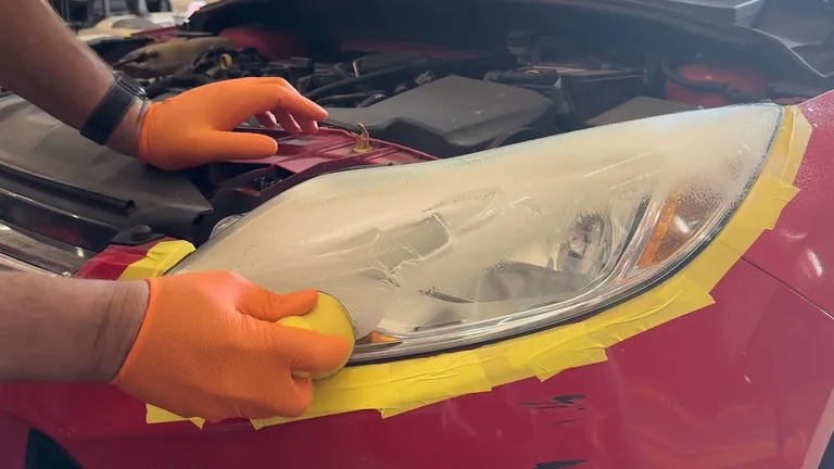 An orange-gloved hand sands the lower headlight lens with a yellow pad while tape covers the surrounding paint.