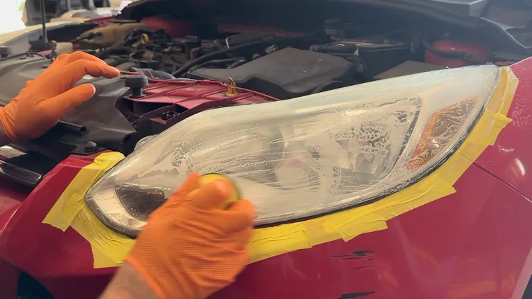 A yellow hand-sanding pad moves across the headlight, with wet sanding residue visible on the lens.